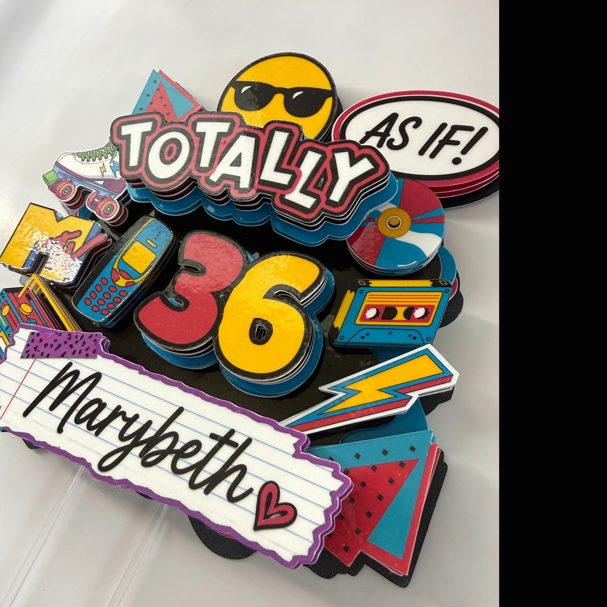 Angled view of a personalized Totally 90s layered birthday cake topper showing dimensional cardstock construction and custom Marybeth name and age 36.