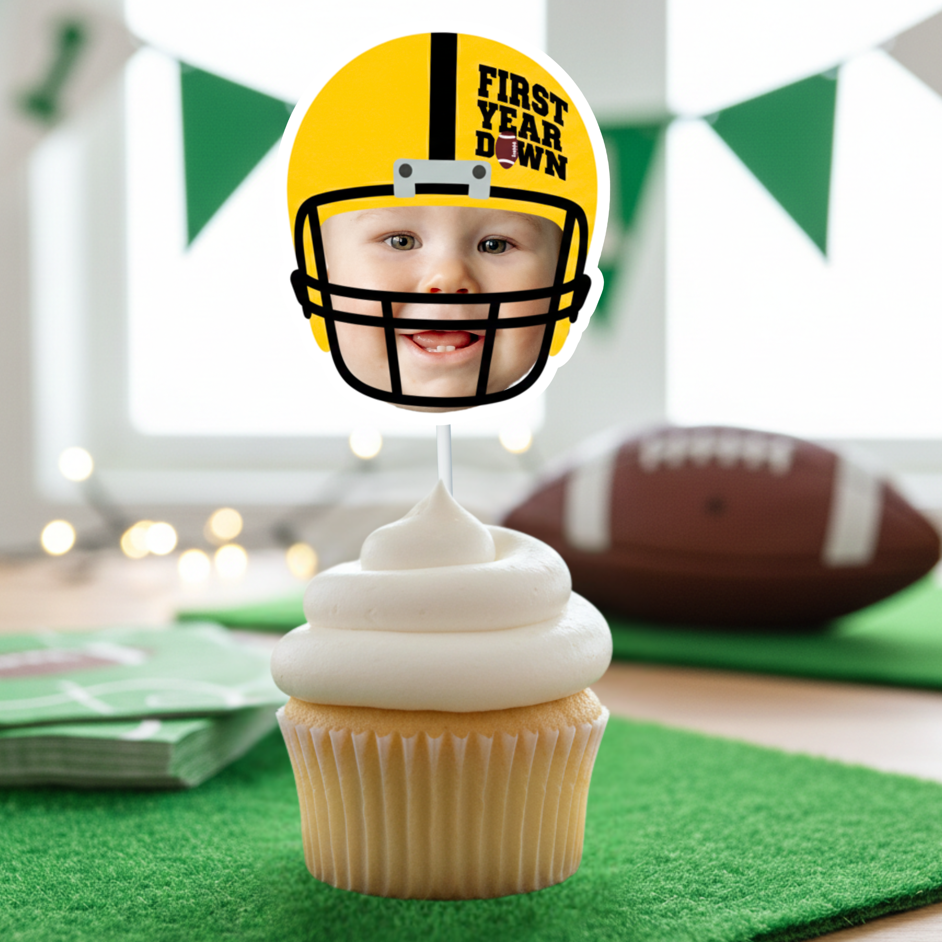 Yellow First Year Down football cupcake topper featuring baby face photo design displayed on vanilla cupcake with white frosting. Styled for football first birthday smash cake celebration with green turf mat, football decor, and sports themed baby boy party background.