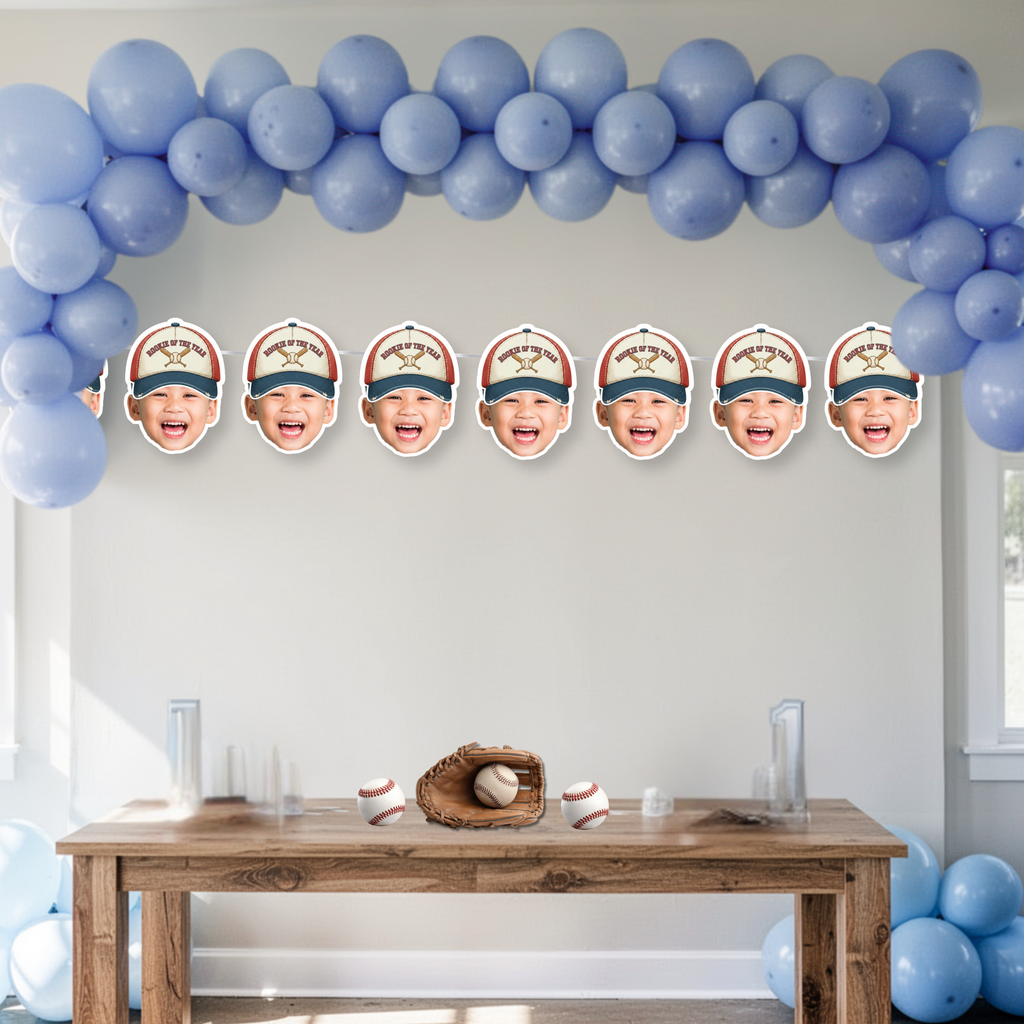Personalized baseball first birthday banner with custom photo faces wearing Rookie of the Year crossed bats hat, styled beneath blue balloon arch as sports themed birthday backdrop and cake smash decor.