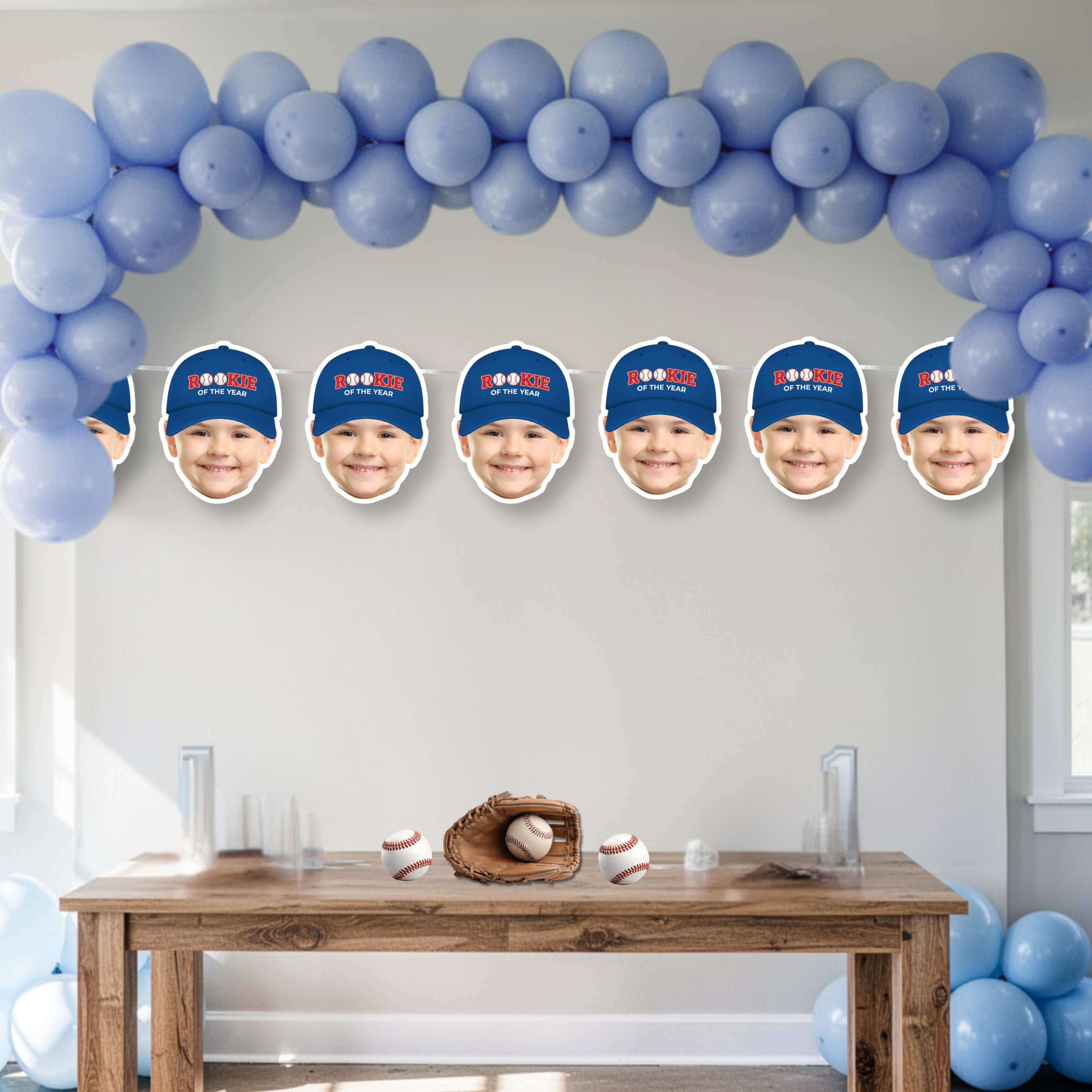 Personalized baseball first birthday banner with custom photo faces wearing blue Rookie of the Year cap, styled beneath blue balloon arch as sports themed birthday backdrop and cake smash decor.