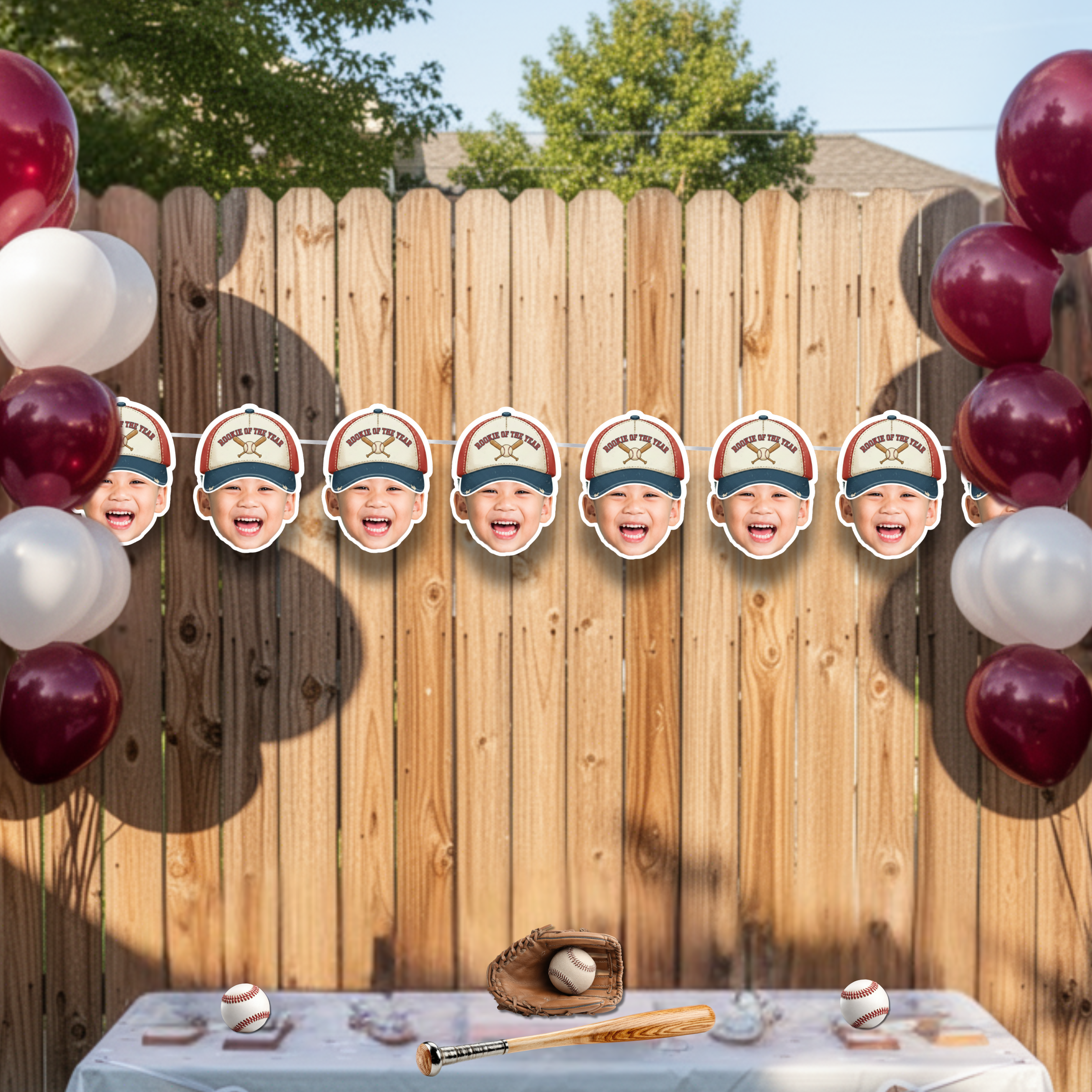 Baseball first birthday photo banner featuring Rookie of the Year crossed bats theme displayed on backyard fence with red and burgundy balloons, styled as outdoor sports birthday party backdrop for little slugger celebration.