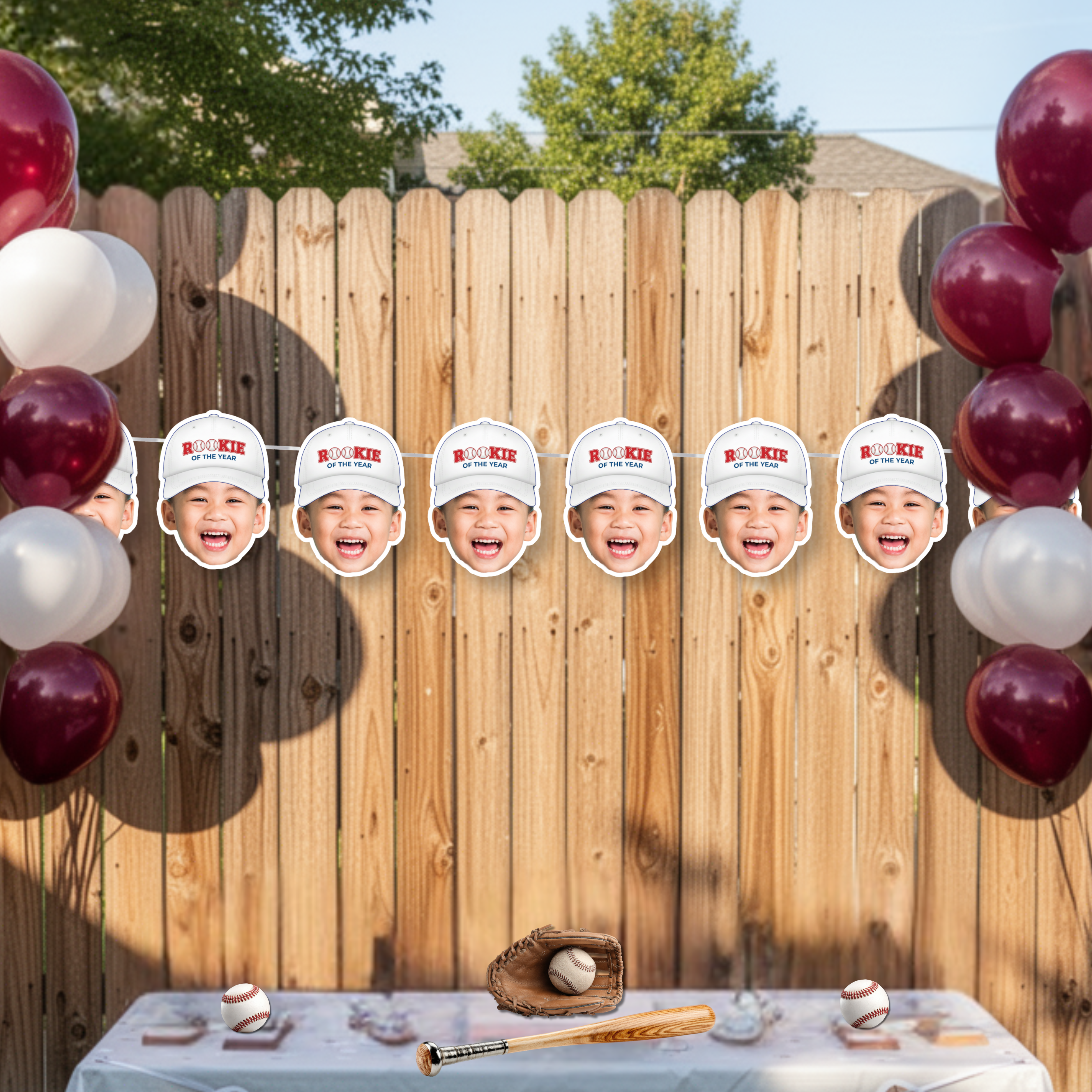 Baseball first birthday photo banner featuring white Rookie of the Year cap design displayed on backyard fence with balloon clusters, styled as patriotic sports birthday party backdrop for little slugger celebration.