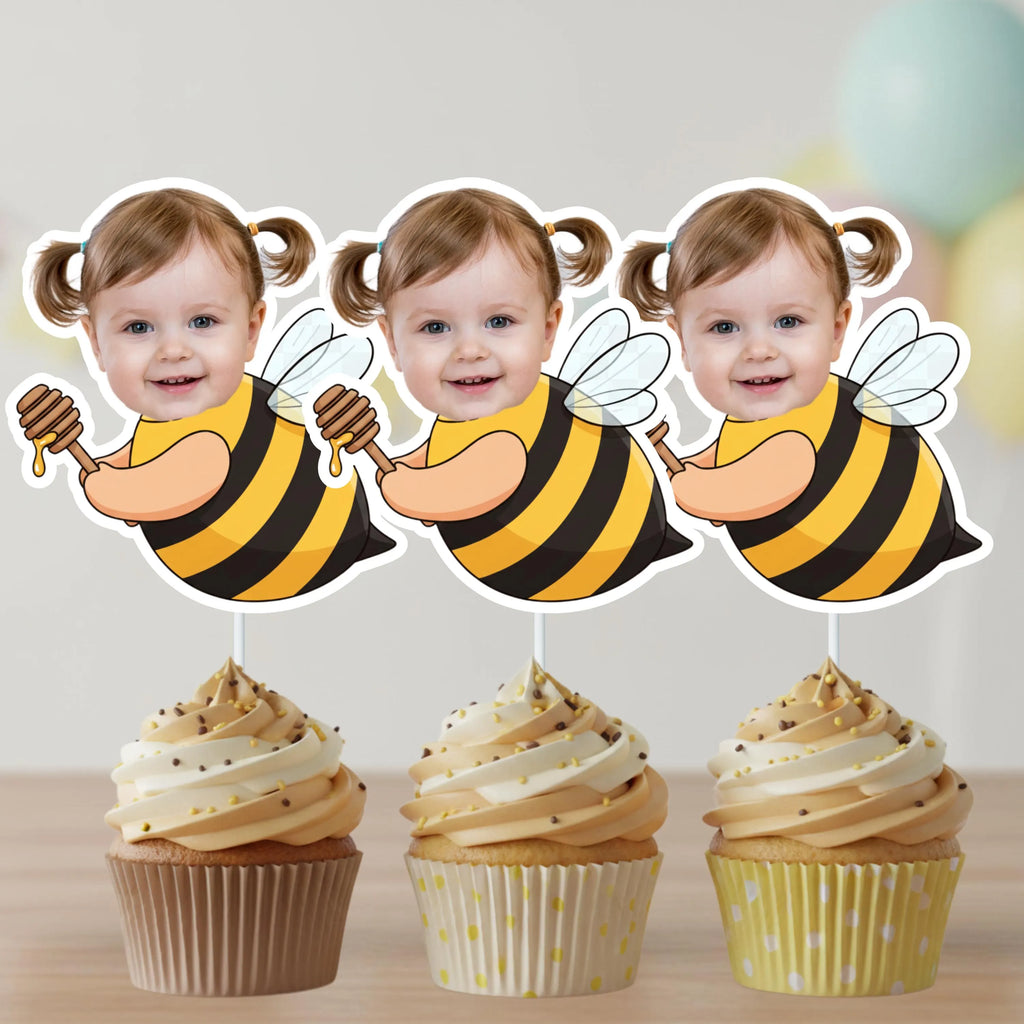 Three cupcakes with bee-themed toppers featuring a child's face, on a light wooden surface.