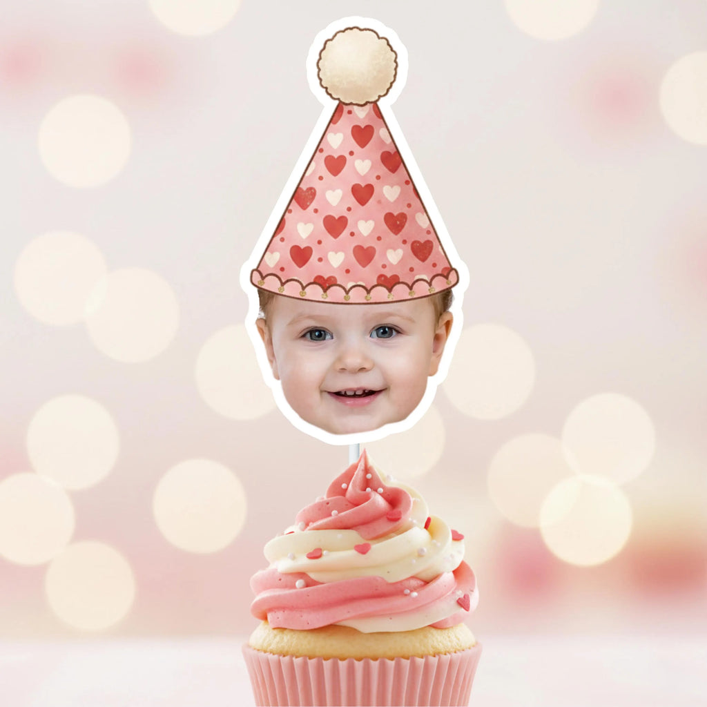 Baby head on a cupcake topper with a heart-patterned party hat against a blurred light background