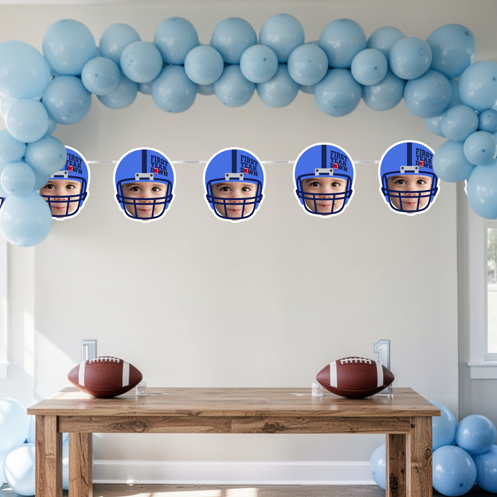 Blue First Year Down football birthday photo banner displayed under a light blue balloon arch. Personalized baby face football helmet banner styled for a boy’s football first birthday party dessert table and smash cake photos.