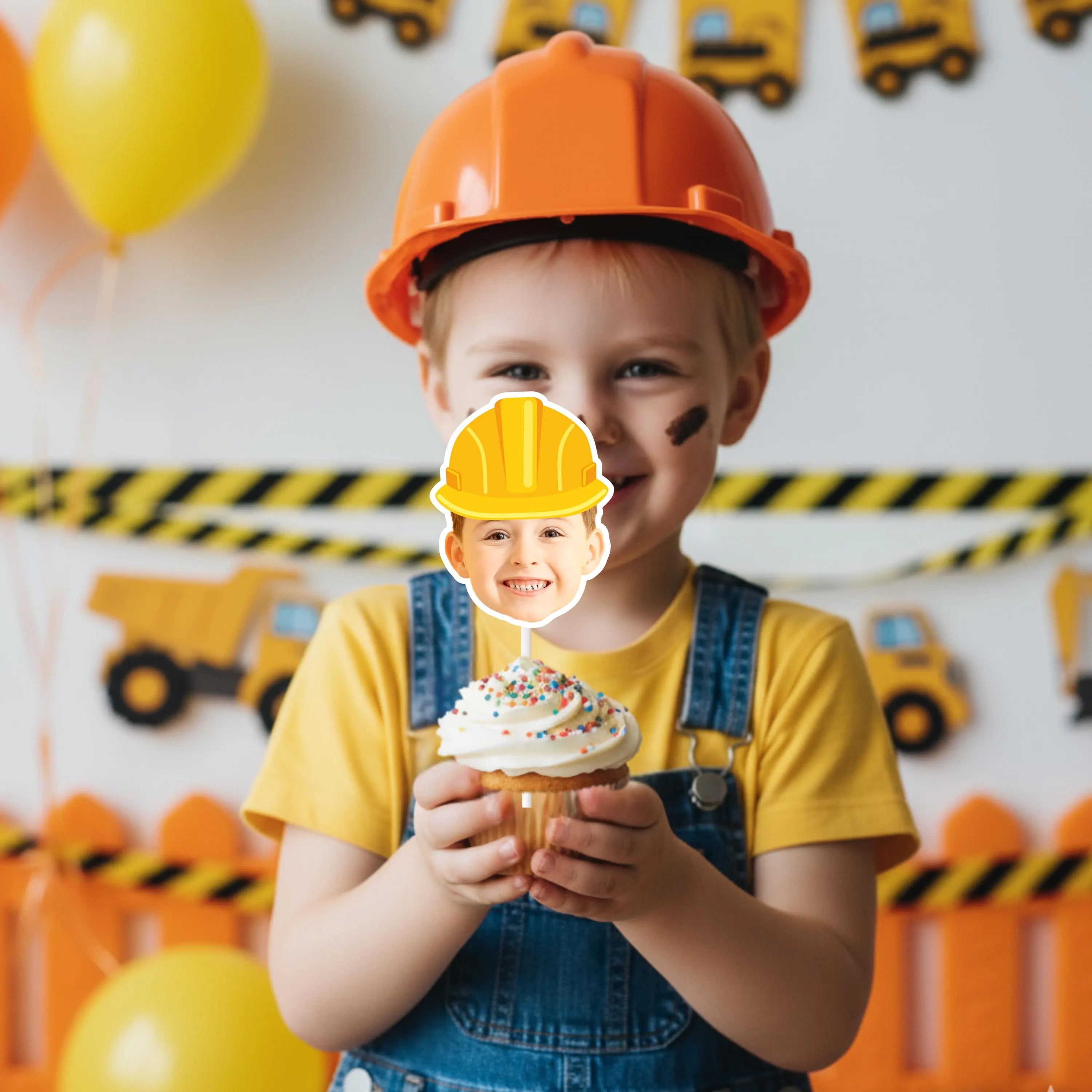 Child in construction-themed outfit holding a cupcake with a face cutout, surrounded by balloons and toy trucks.