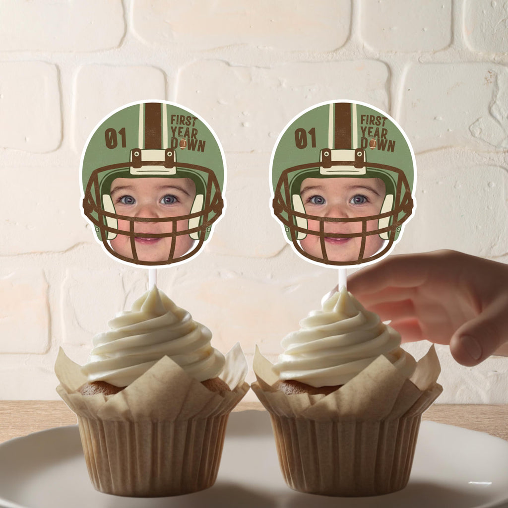 Two cupcakes with personalized toppers featuring a baby’s face inside a vintage green football helmet with 'First Year Down' text.