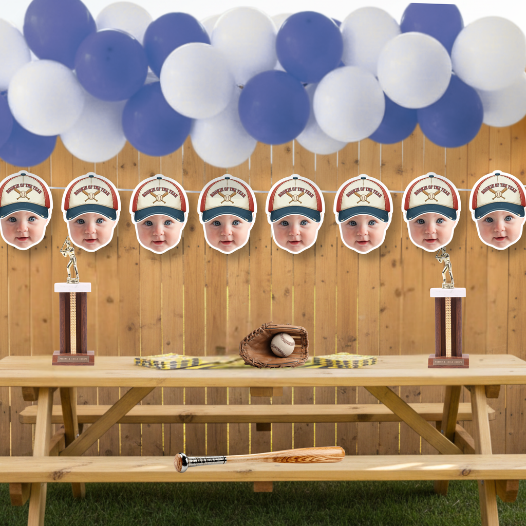 Custom photo baseball banner for Rookie of the Year party displayed on wooden backyard fence with blue and white balloon garland, baseball trophies, glove and bat decor for outdoor little slugger birthday celebration.