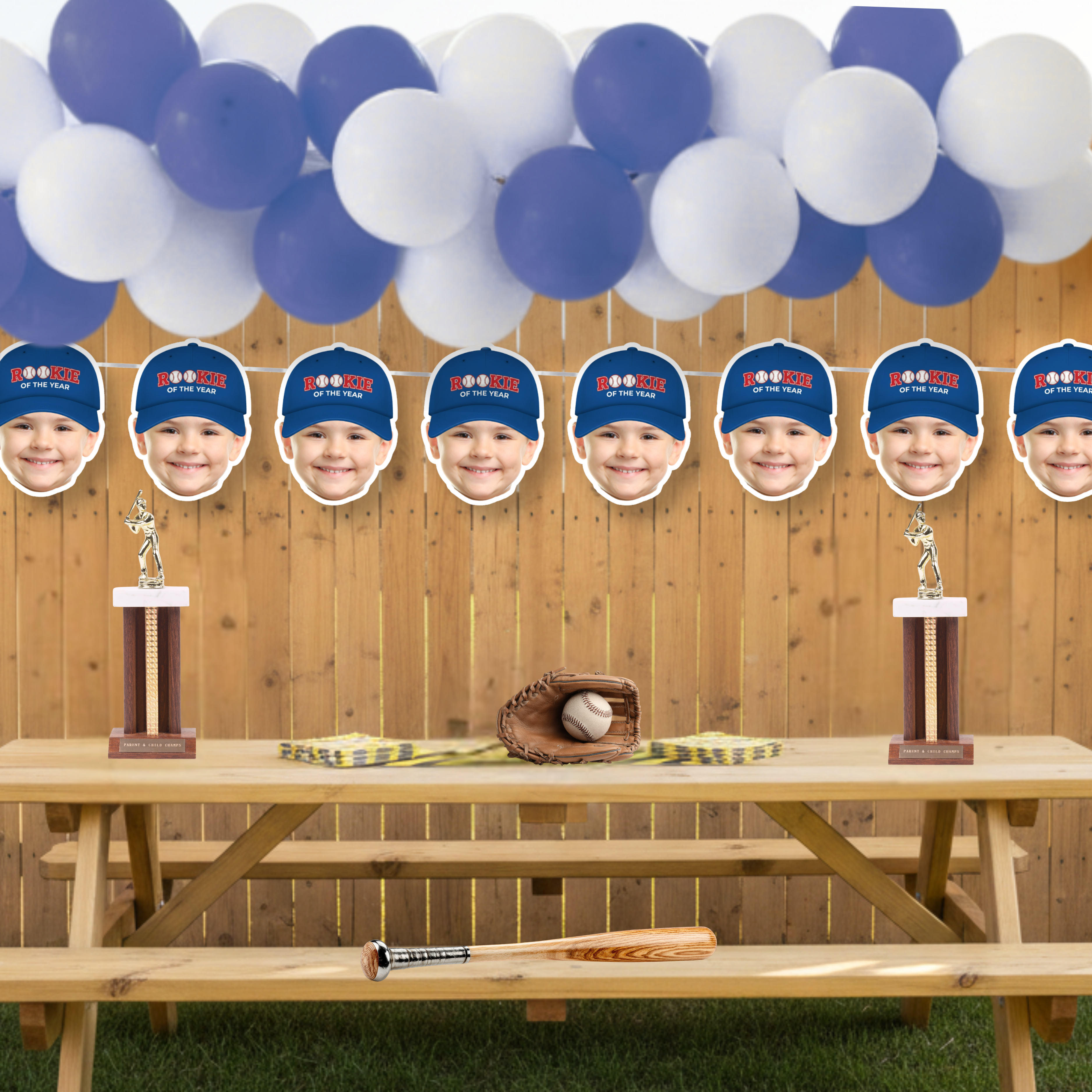 Custom photo baseball banner in blue Rookie of the Year design displayed on backyard wooden fence with balloon garland, baseball trophies, glove and bat decor for outdoor little slugger birthday celebration