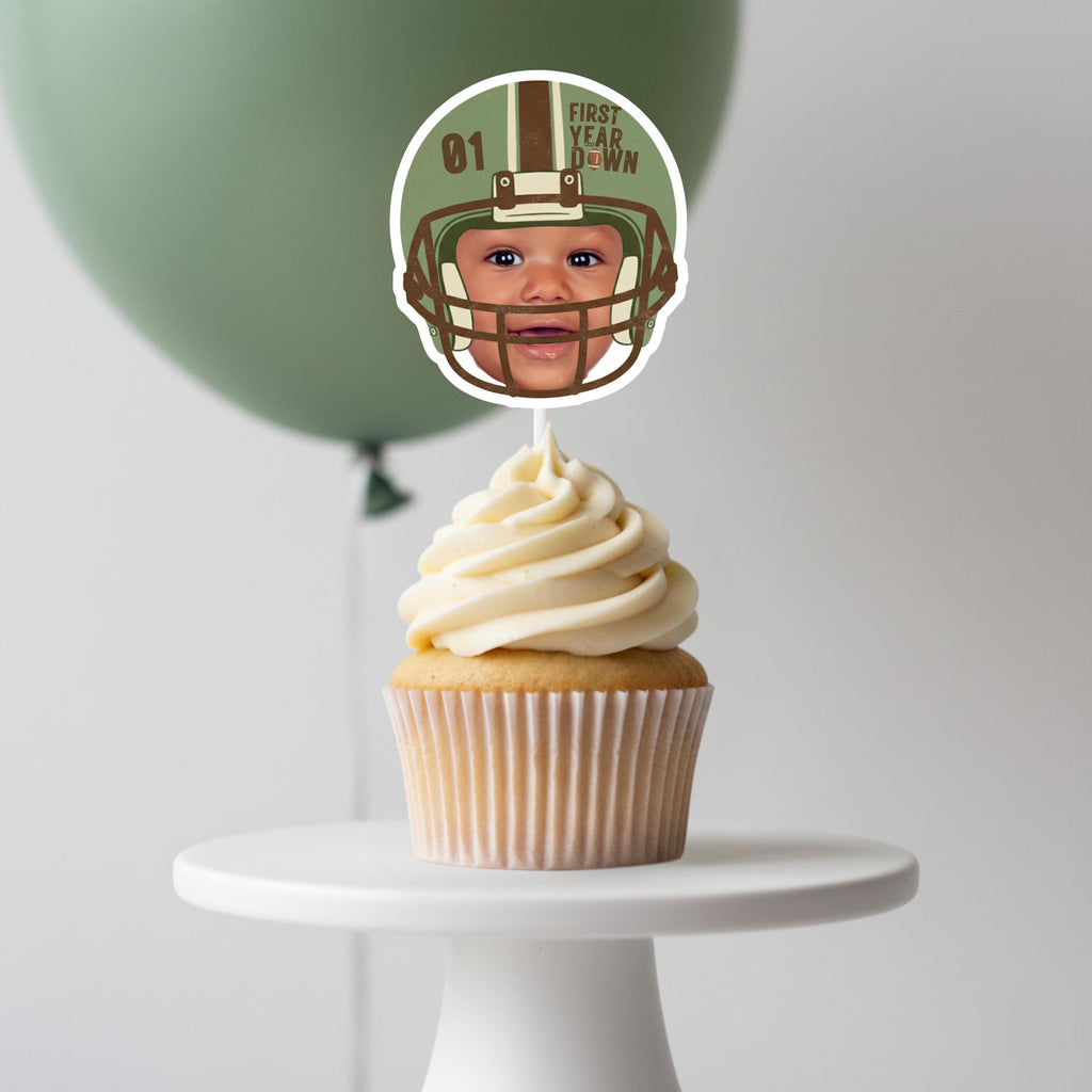 A single cupcake on a white stand featuring a high-quality personalized baby face topper in a green football helmet for a 1st birthday celebration.