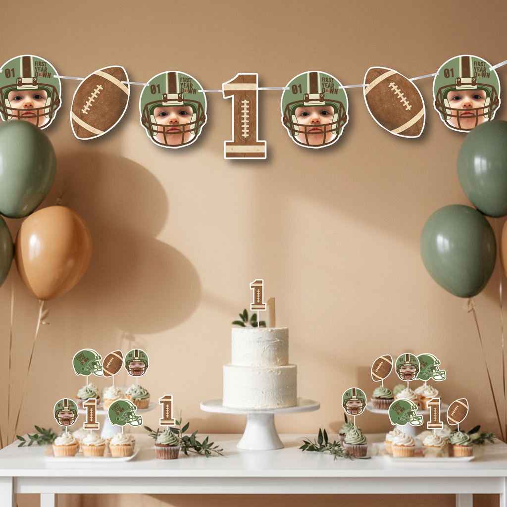 First Year Down football first birthday banner styled above a neutral dessert table with smash cake, cupcakes, and sage green and tan balloons. Custom photo football banner decor designed for a sports themed one year old birthday party celebration.