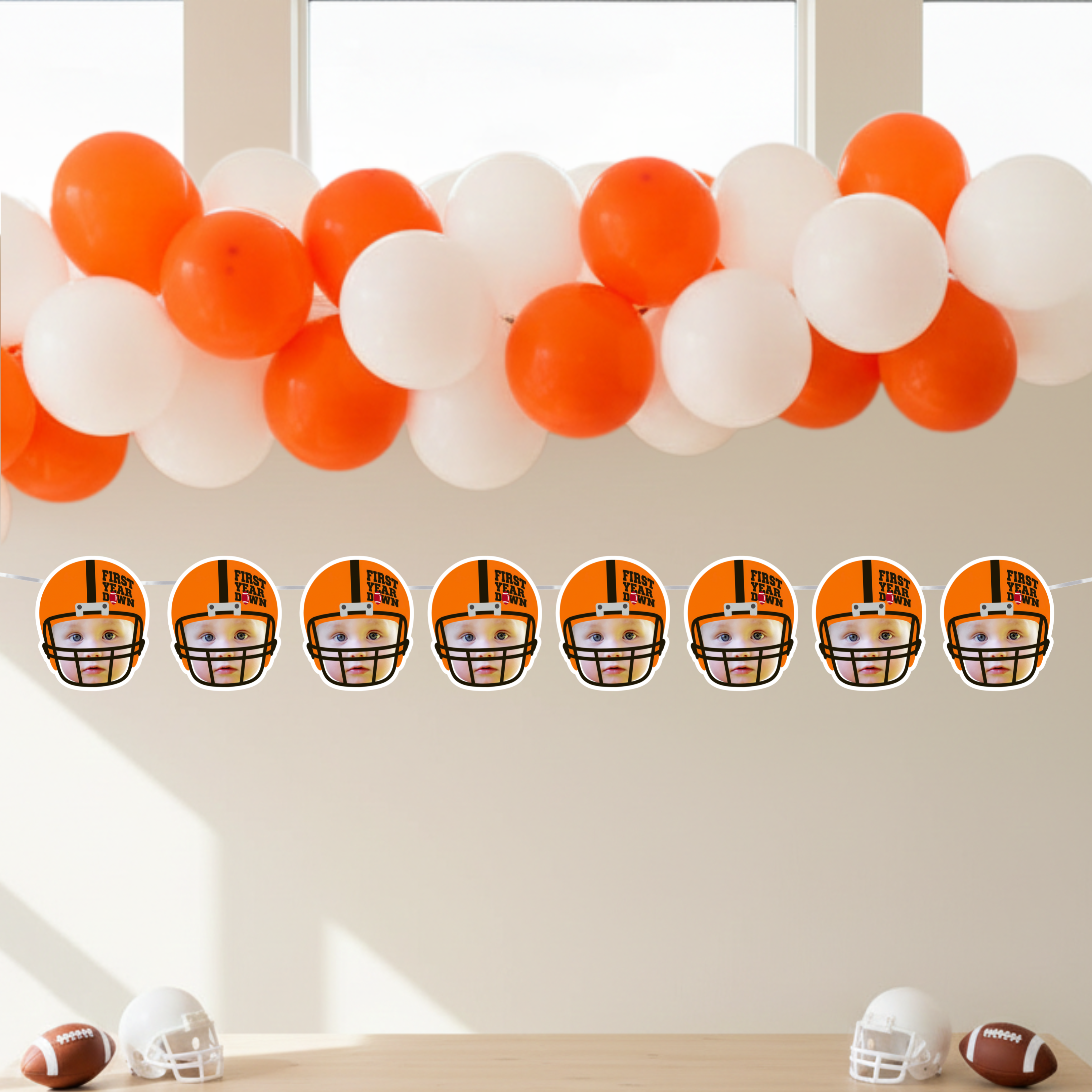 First Year Down football birthday photo banner displayed under an orange and white balloon garland for a boy’s first birthday party. Personalized football helmet face banner strung across a modern dessert table setup for a Rookie of the Year football theme celebration.