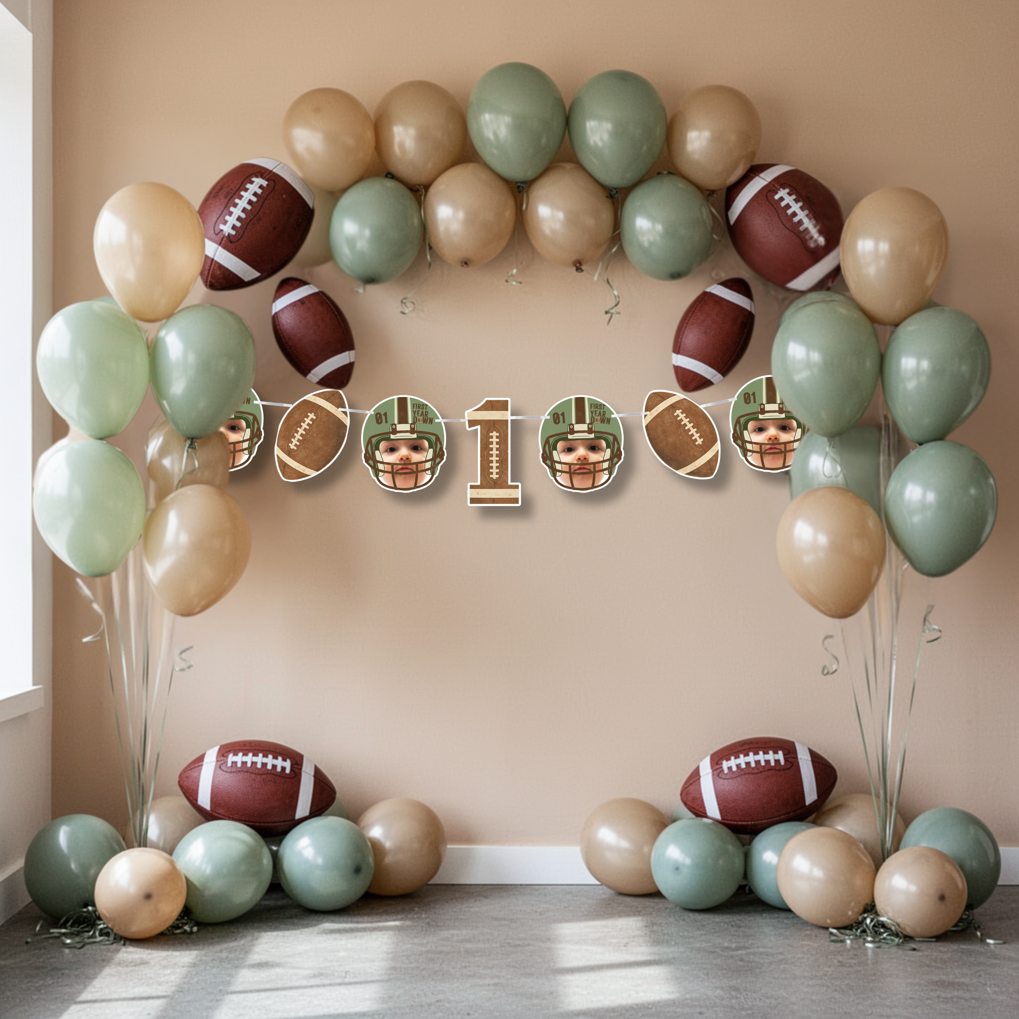 Football first birthday party backdrop featuring a large balloon arch in sage green and tan with First Year Down banner centered underneath. Sports themed one year old celebration decor with football accents and custom baby photo elements.