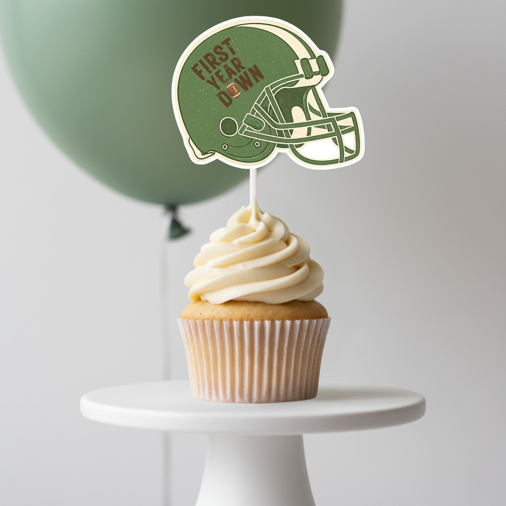 Green football helmet cupcake topper with First Year Down lettering displayed on a frosted cupcake with soft green balloon backdrop. Perfect accent for football first birthday party supplies and game day inspired baby celebration décor.