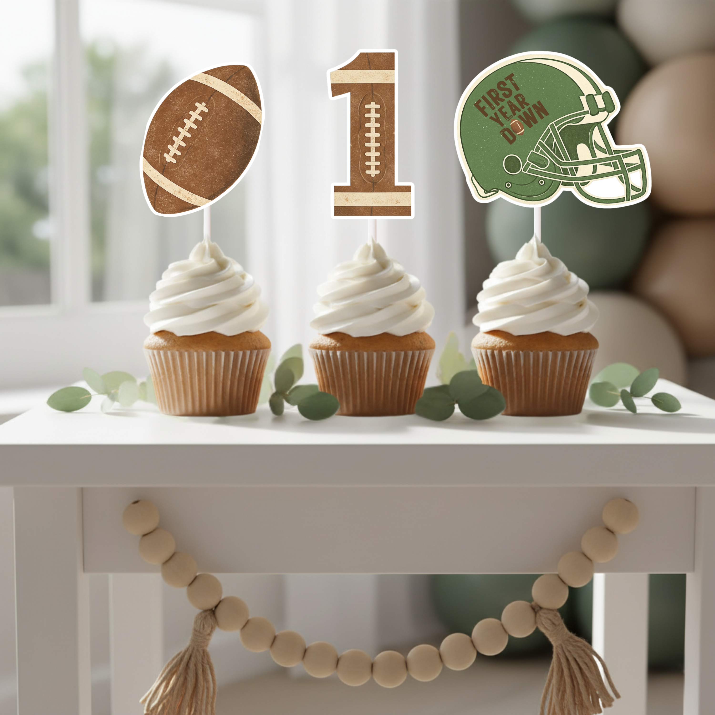 Football First Year Down cupcake toppers displayed on vanilla frosted cupcakes on a white dessert table with wood bead garland. The set includes a vintage brown football, textured number one, and green helmet with First Year Down design, perfect for a rustic football themed first birthday party.