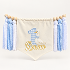 Baby blue One Silly Goose personalized first birthday high chair banner with custom name and age, cream canvas pennant, natural wooden beads, and blue and white yarn tassels.