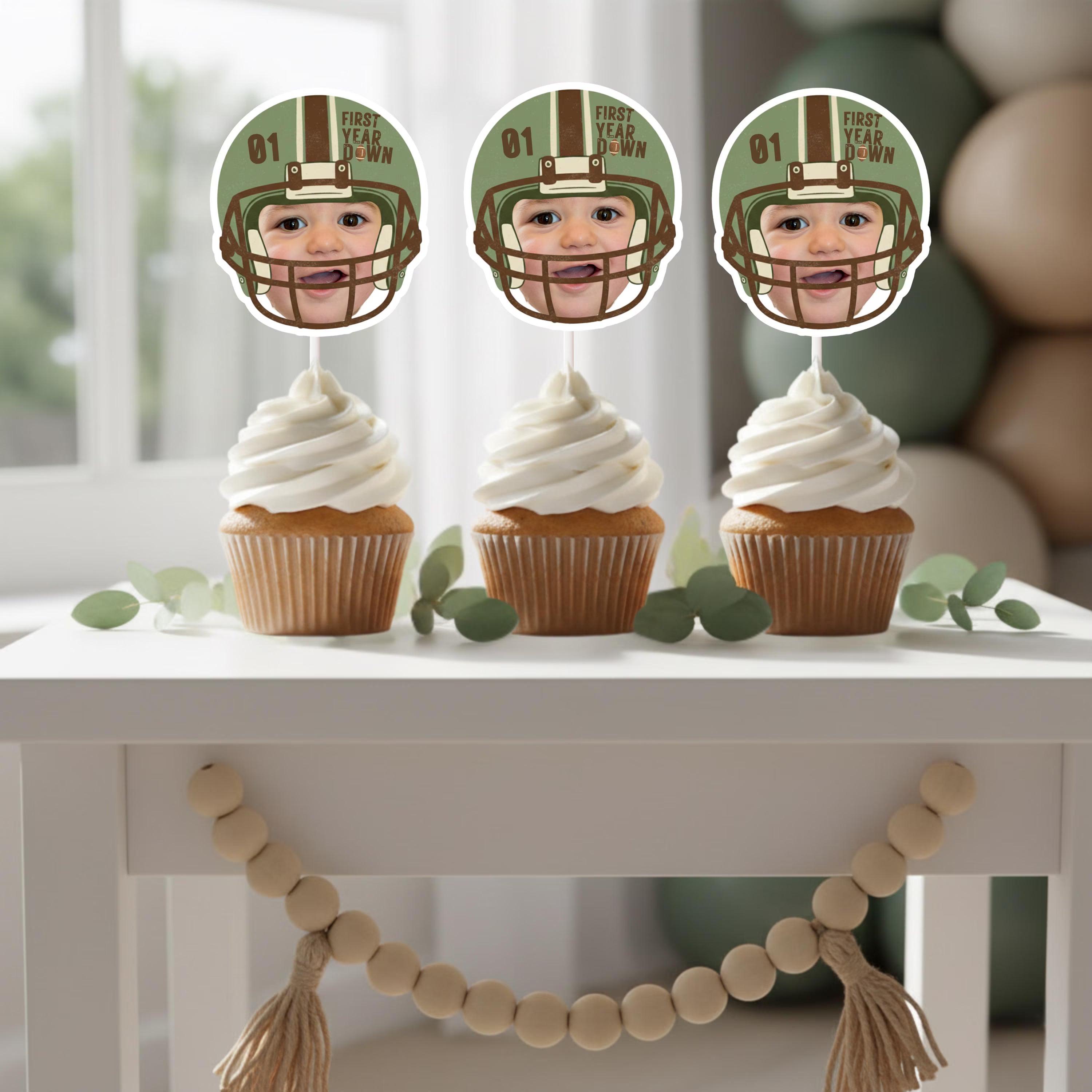 Three cupcakes with custom baby face photo toppers arranged on a white table with eucalyptus and a wood bead garland for a modern football party.