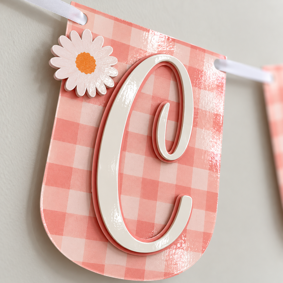 Close-up of a laminated pink gingham banner panel with a layered white script letter and raised daisy detail.