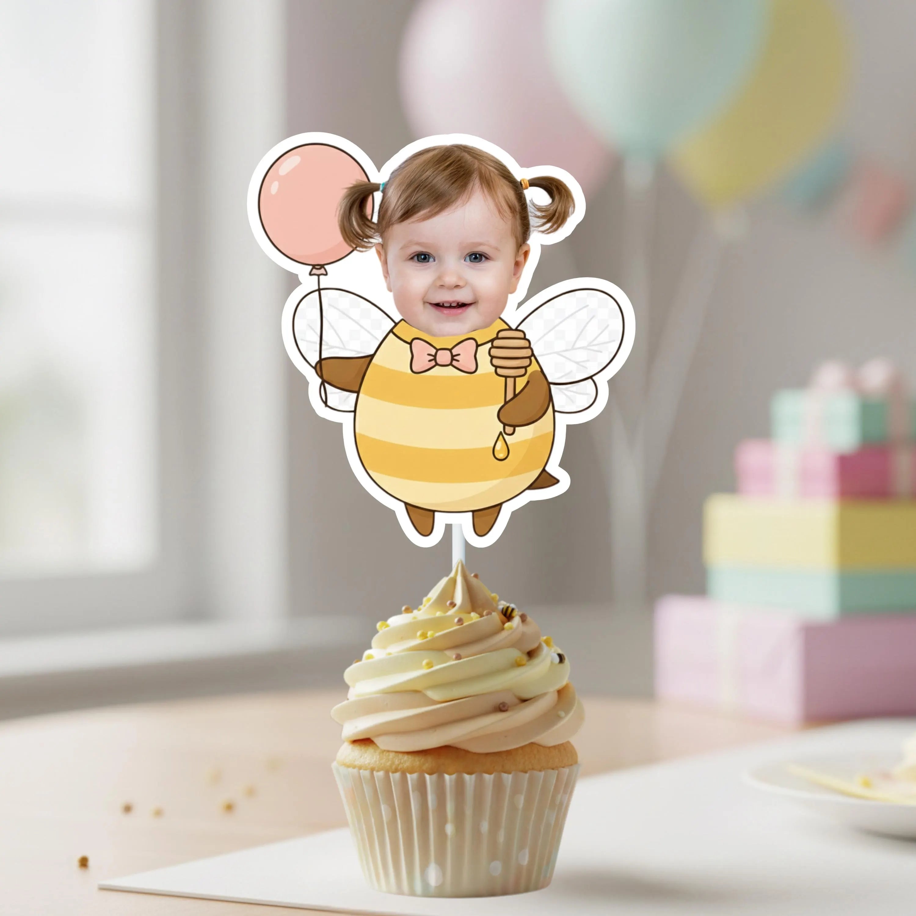 Cupcake with a bee-themed topper featuring a child's face, balloons in the background.