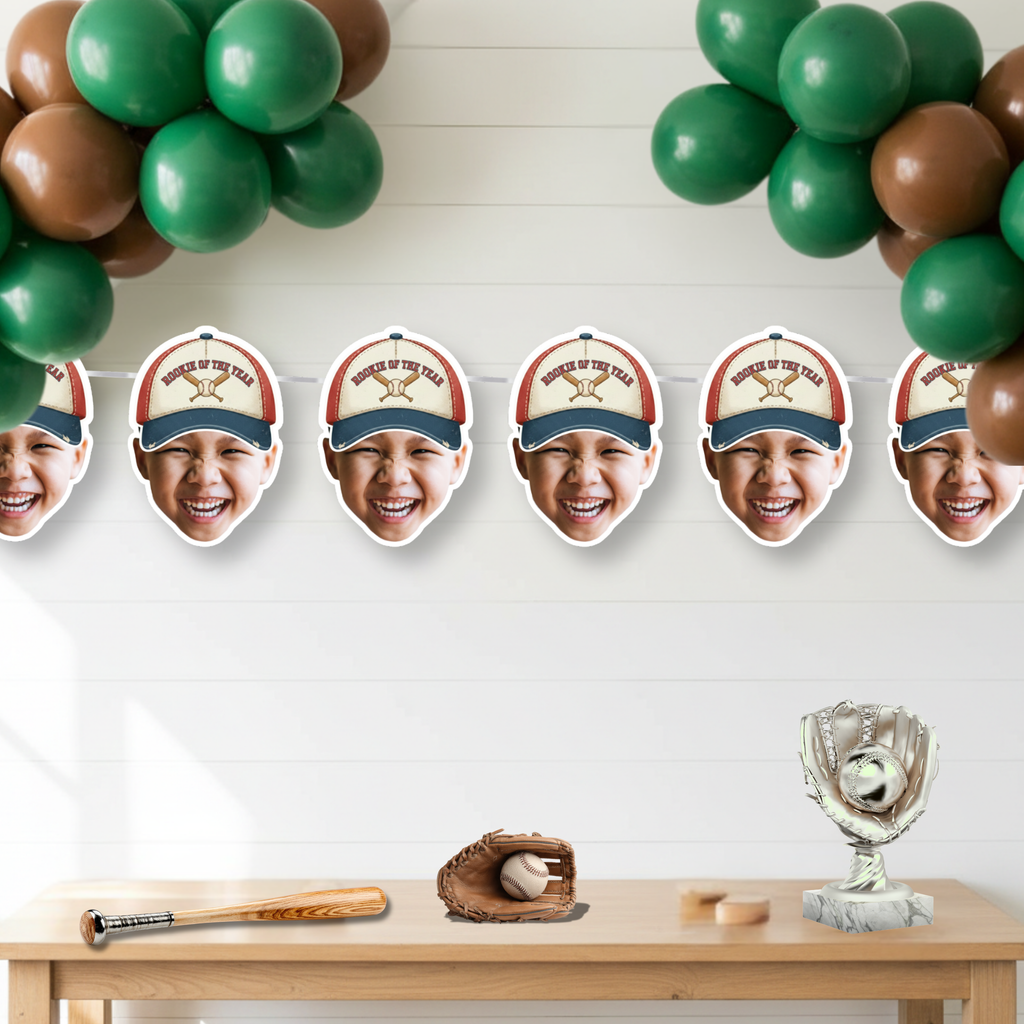 Photo baseball Rookie of the Year banner featuring crossed bats cap design displayed under green and brown balloon garland above dessert table with baseball bat, glove, and trophy decor for a baseball first birthday party.