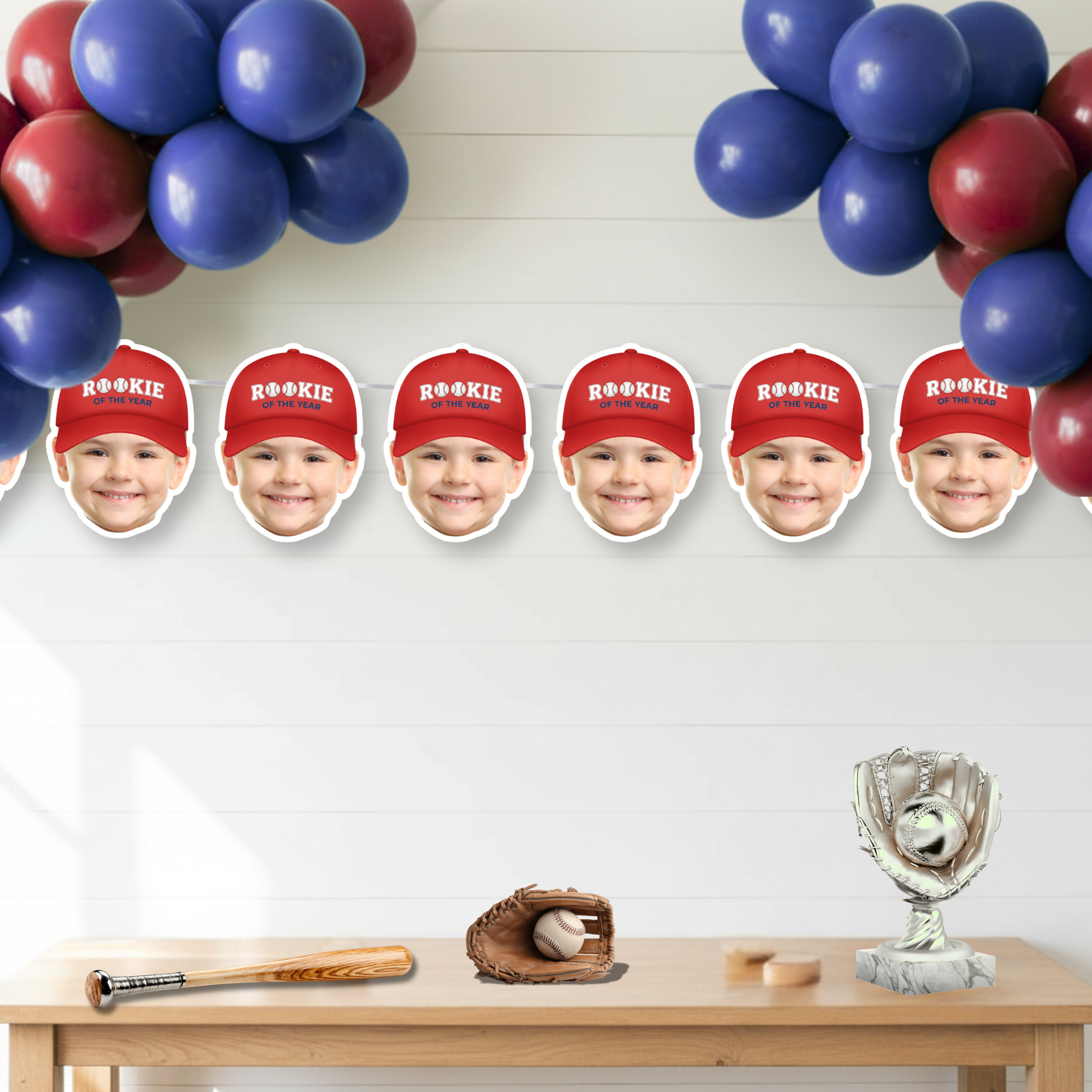 Photo baseball Rookie of the Year banner featuring red cap design displayed under red and blue balloon garland above dessert table with baseball bat, glove, and trophy decor for a baseball first birthday party.