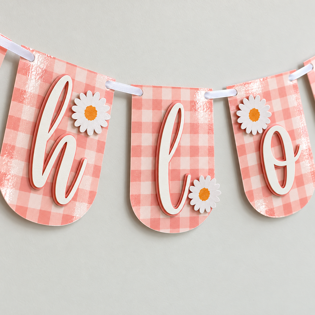 Close-up of personalized pink gingham birthday banner panels showing raised script letters, layered daisies, lamination, and white satin ribbon.